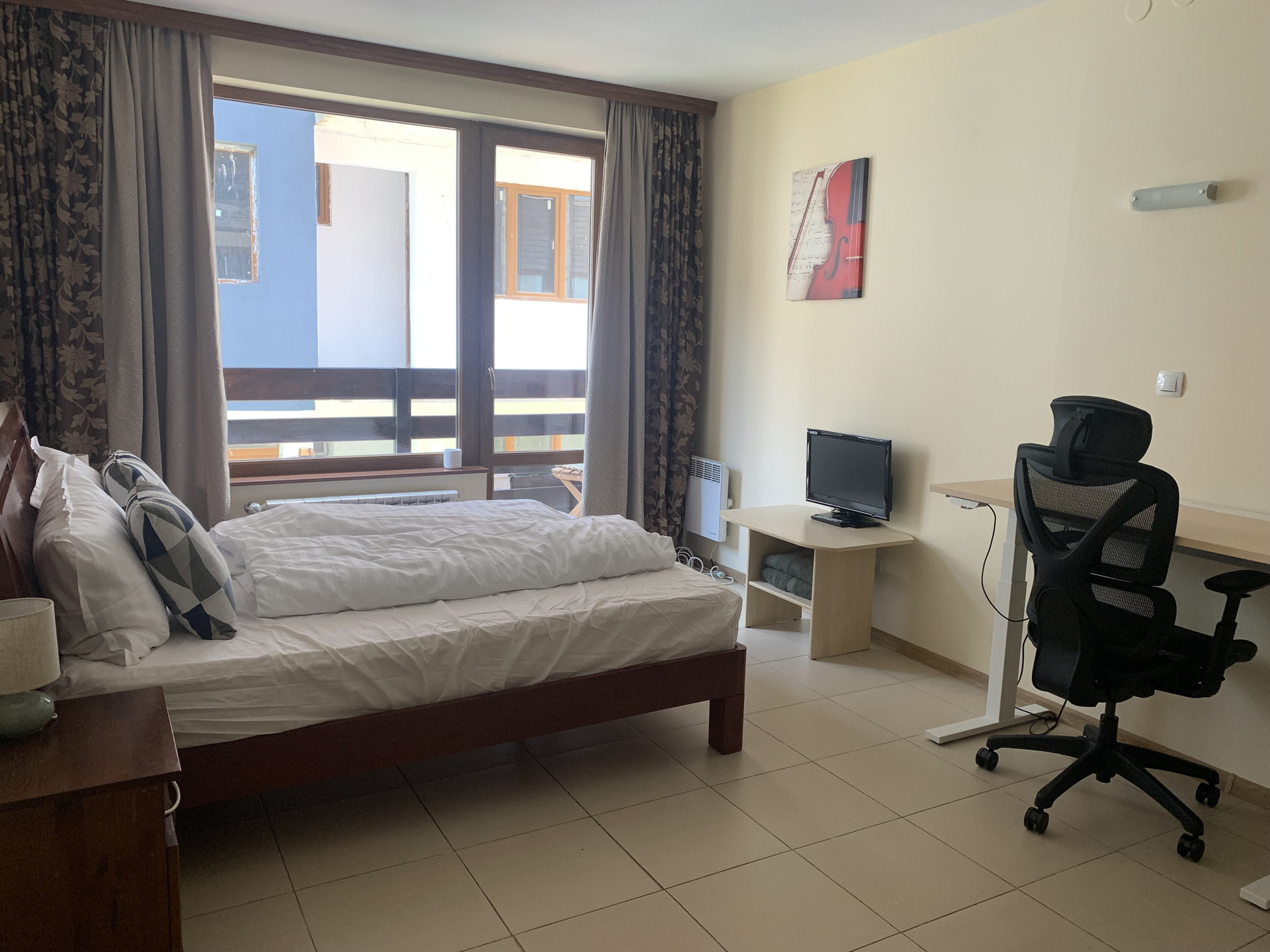 Bright studio overview: double bed, balcony window with mountain/courtyard view, ergonomic desk, chair and TV.