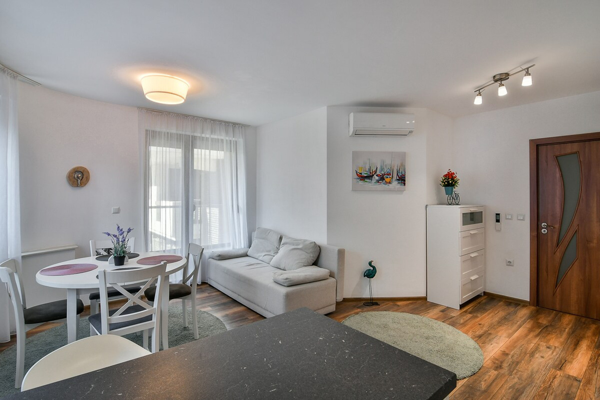 Bright and stylish open-plan living space featuring a cozy sofa, elegant dining area, and modern decor. Natural light fills the room through large windows, creating a warm and inviting atmosphere. Perfect for relaxing, dining, or socializing