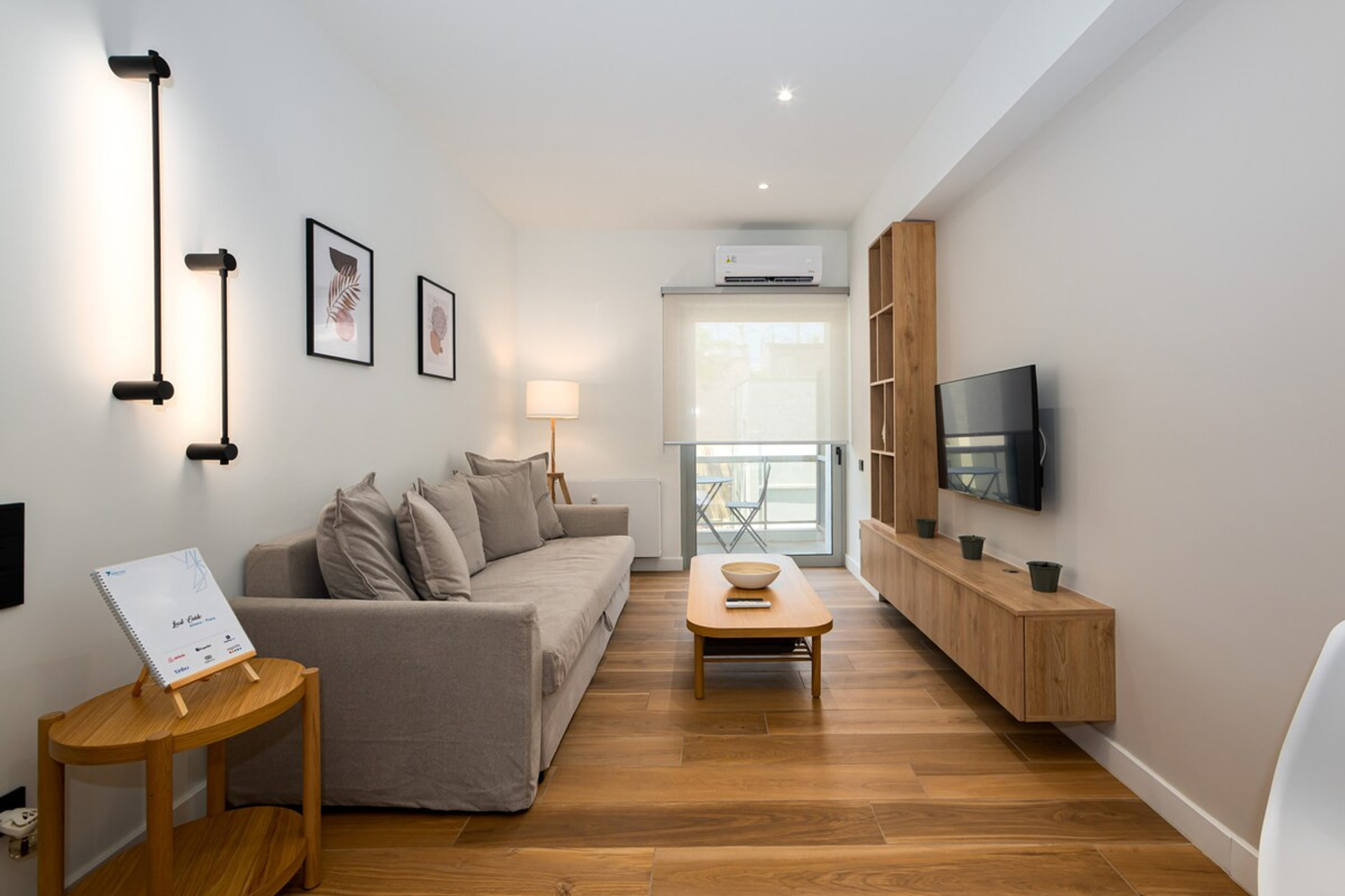 A bright, modern living space featuring a cosy grey sofa, stylish wooden furniture, and elegant wall lighting. The room is complemented by warm wooden flooring, tasteful decor, and a large TV, creating a welcoming and relaxing ambiance.