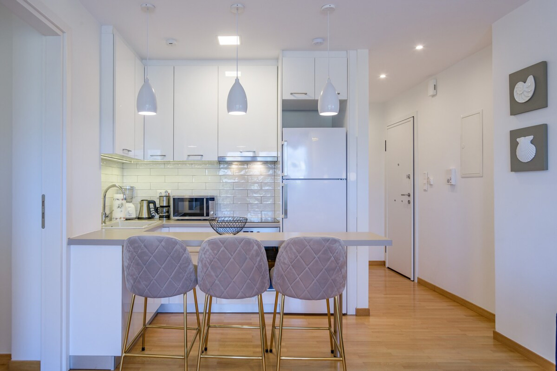 Bright and modern kitchen with sleek white cabinetry, stylish pendant lighting, and a cosy breakfast bar featuring plush seating. Perfectly equipped for your cooking needs, this inviting space blends functionality with elegance for a delightful stay.