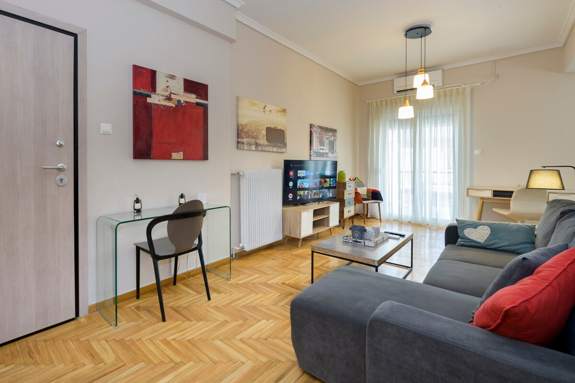 Bright and inviting living room with modern decor, featuring a cosy sofa, smart TV, and a sleek glass desk for work or leisure. Natural light floods through large windows, complemented by warm wooden flooring and tasteful artwork for a homely touch.