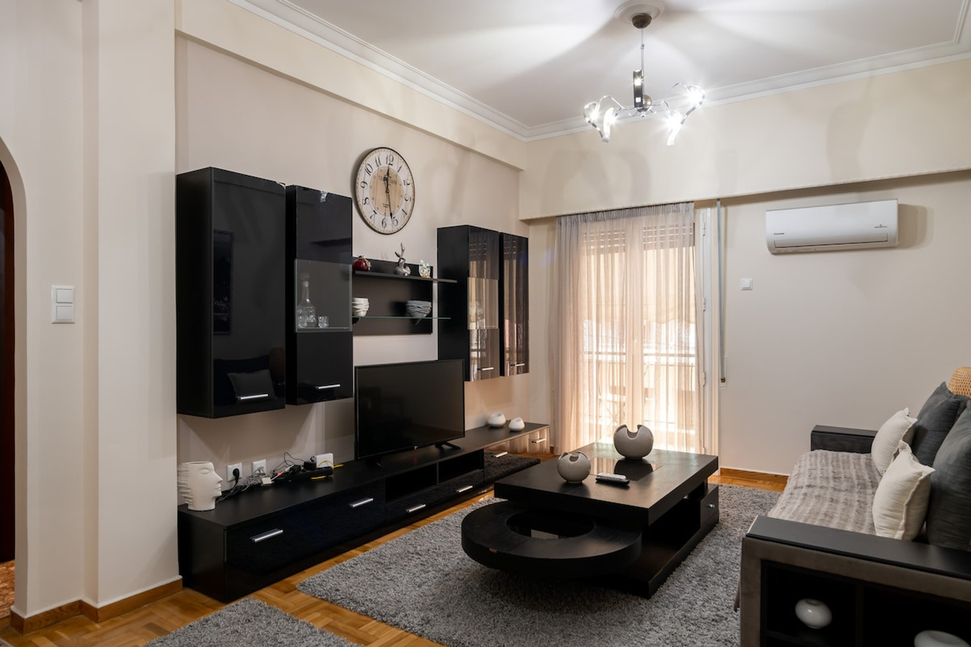 A stylish and spacious living room featuring sleek black furniture, a cosy sofa, and a modern chandelier. Natural light flows through large windows with sheer curtains, creating a warm and inviting atmosphere perfect for relaxation or entertaining.