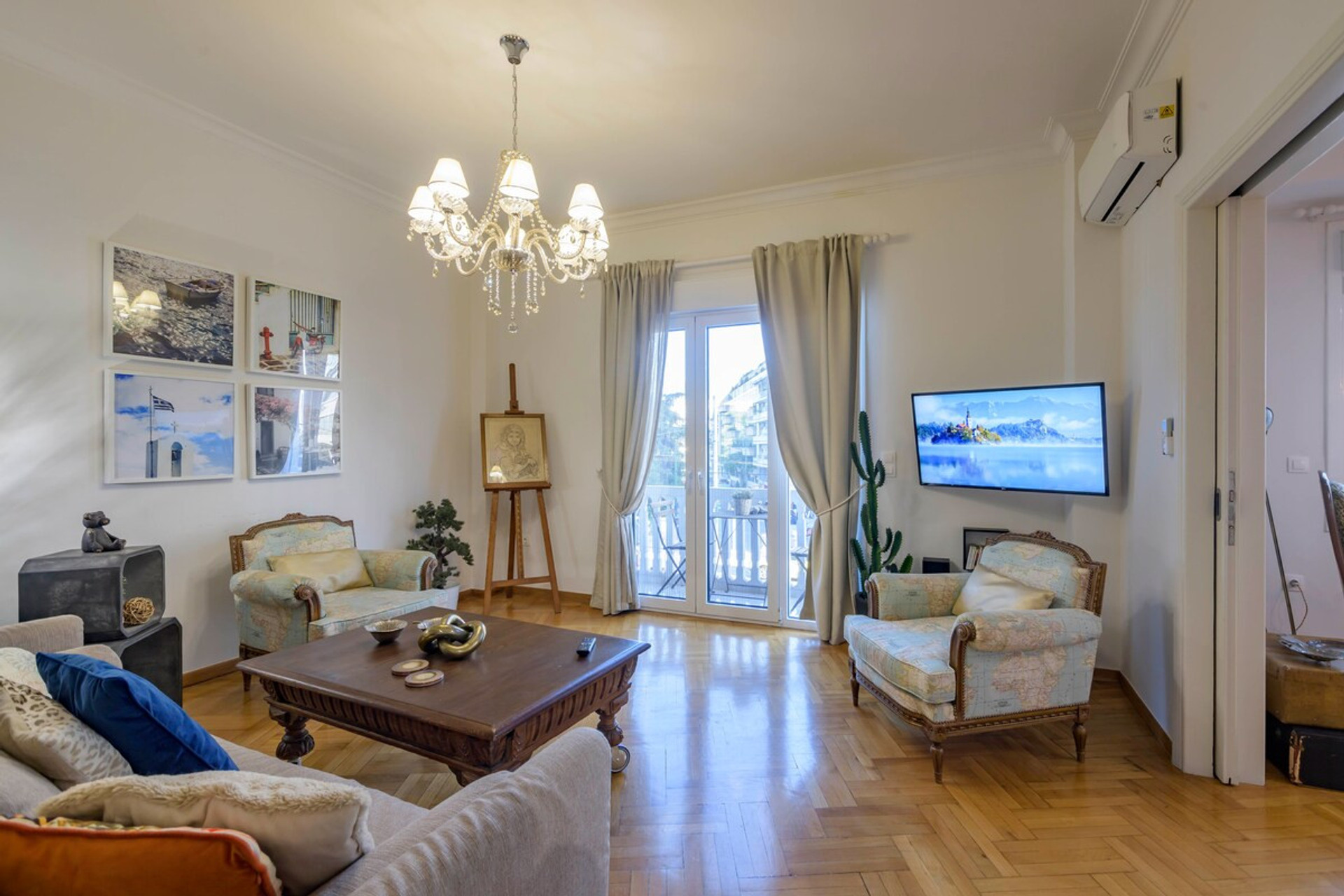 Bright and elegant living room featuring a cosy sofa, stylish armchairs, and a charming chandelier. Enjoy natural light through French doors leading to a balcony, complemented by tasteful decor and a large smart TV for your entertainment.