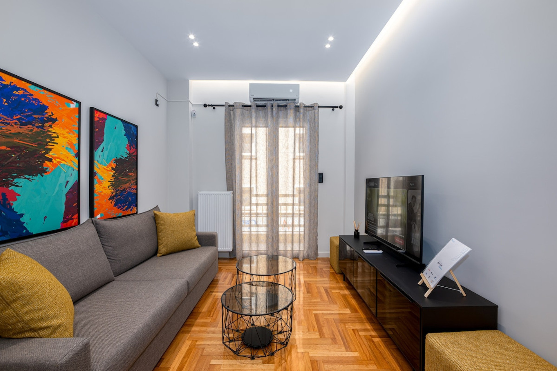 Bright and modern living space with vibrant artwork, cosy seating, and warm wooden flooring. The room is complemented by a sleek TV unit, soft lighting, and natural light streaming through elegant curtains, creating a perfect spot to relax and unwind
