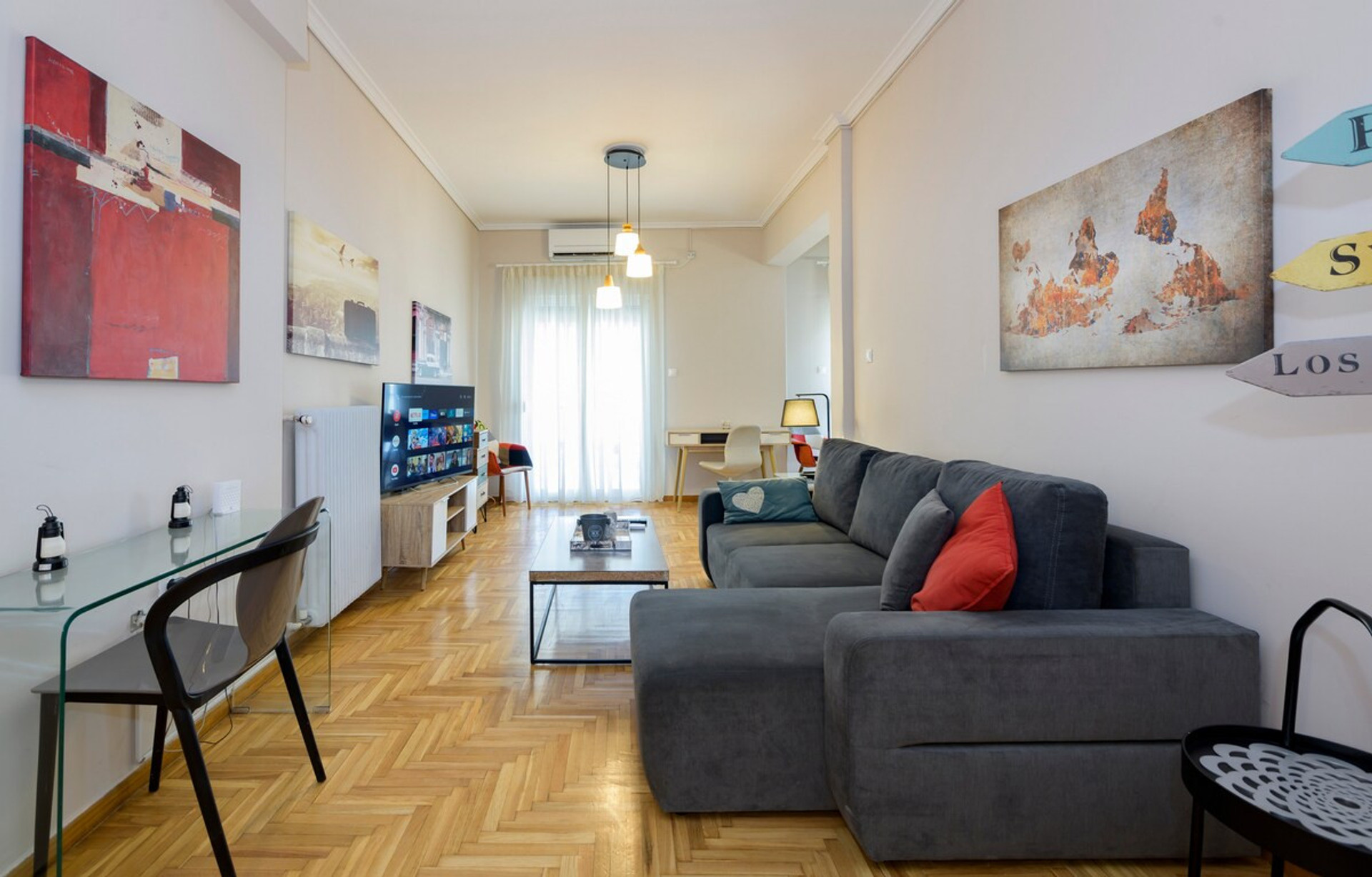 A bright and inviting living room featuring a cosy grey sofa adorned with colourful cushions, a sleek glass desk, and vibrant artwork on the walls. The space is complemented by natural light streaming through large windows, creating a warm atmosphere