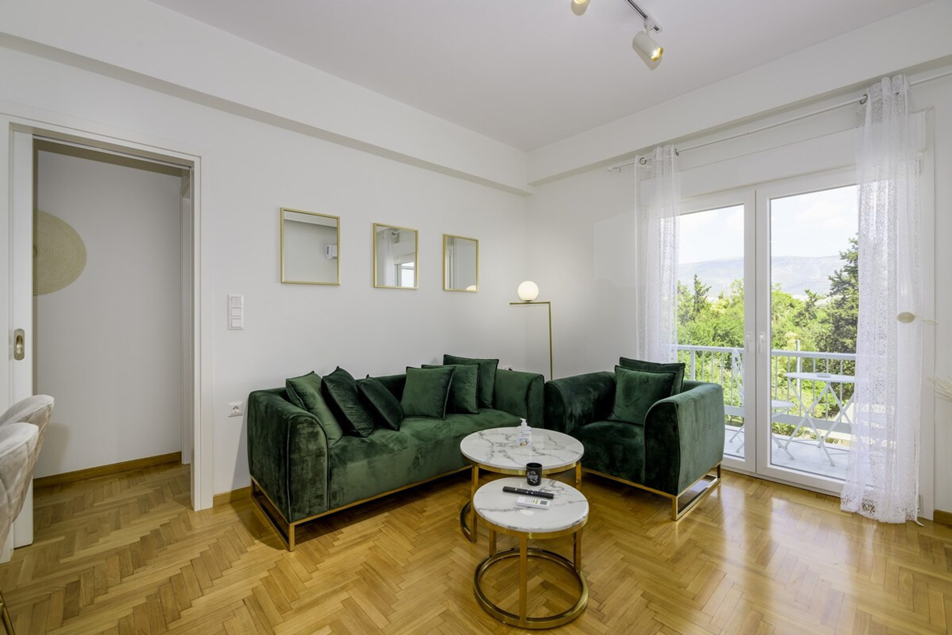 Bright and stylish living room featuring a plush green sofa, elegant marble coffee tables, and floor-to-ceiling windows opening to a serene balcony with lush views. Perfect for relaxing or entertaining in comfort and style. 