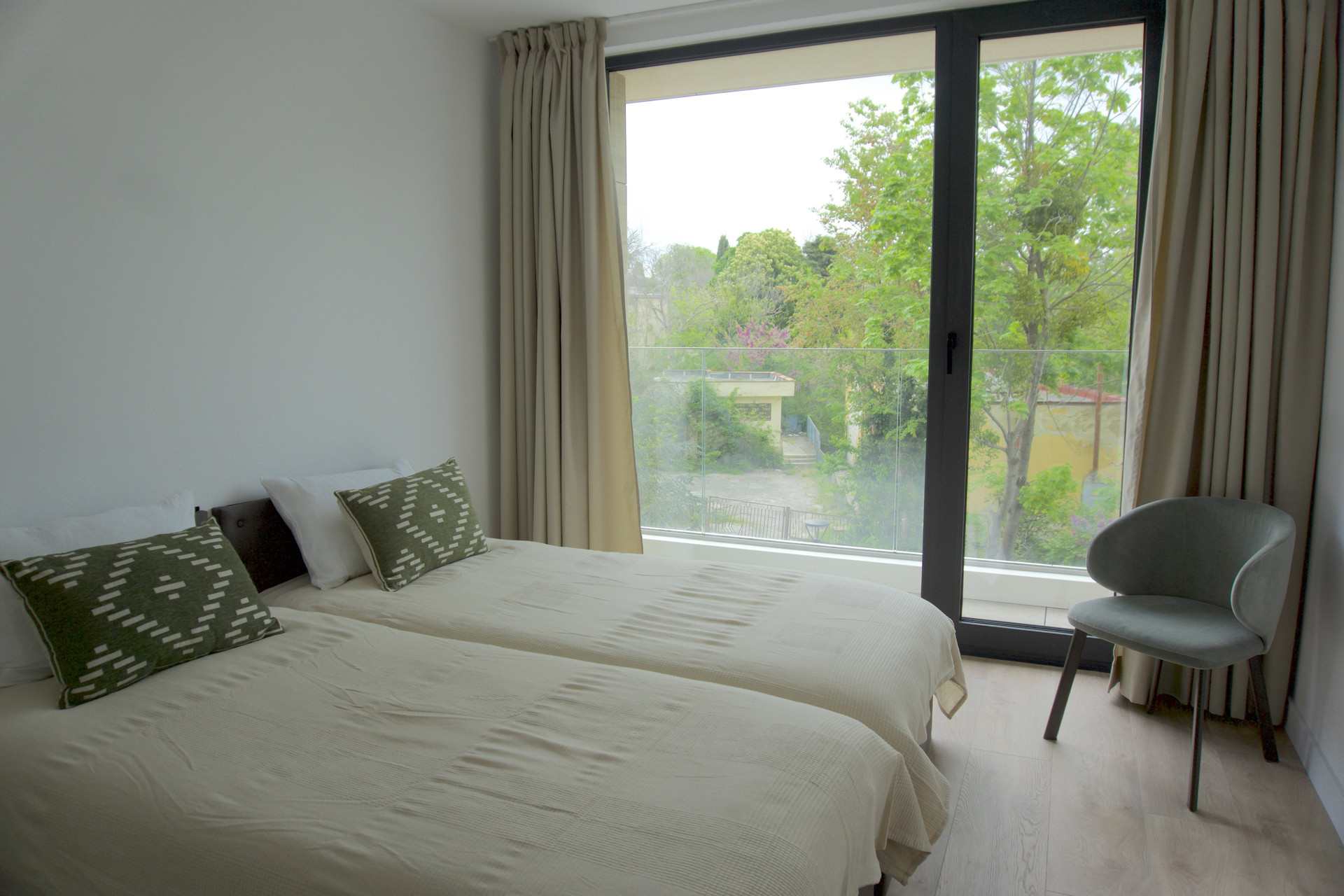 Wake up to stunning, uninterrupted nature views through the expansive floor-to-ceiling windows. This bright, tranquil bedroom serves as your personal sanctuary. It features floor-length curtains for a perfect night's rest, comfortable twin beds that can be configured to suit your needs, and a cozy modern accent chair that is perfect for reading or simply taking in the lush green surroundings.