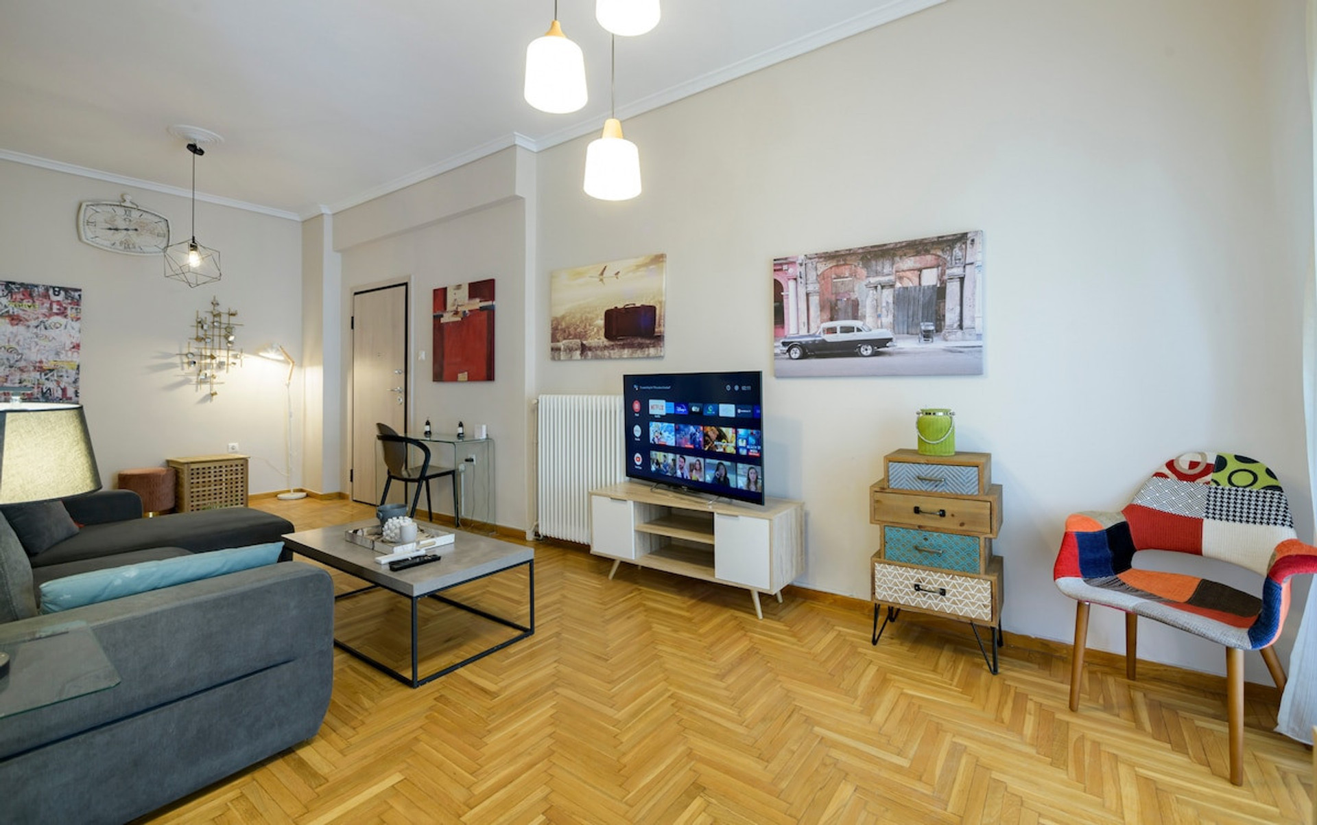A bright and inviting living room featuring a cosy sofa, stylish décor, and a smart TV for entertainment. The space is enhanced by warm lighting, parquet flooring, and a colourful accent chair, creating a perfect setting to relax and unwind.