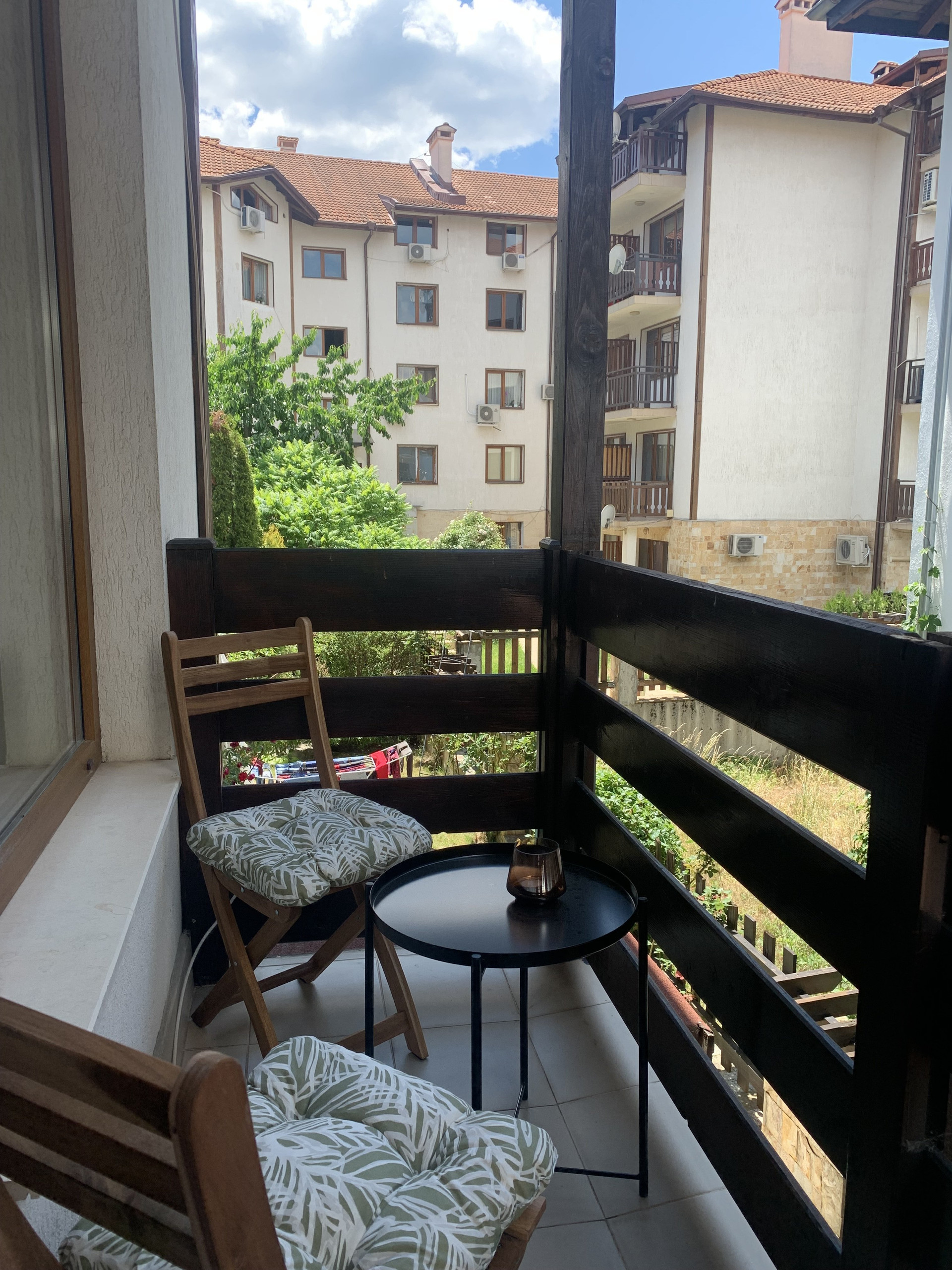 Private balcony under a wooden pergola with cushioned chairs and a side table, overlooking the green courtyard.