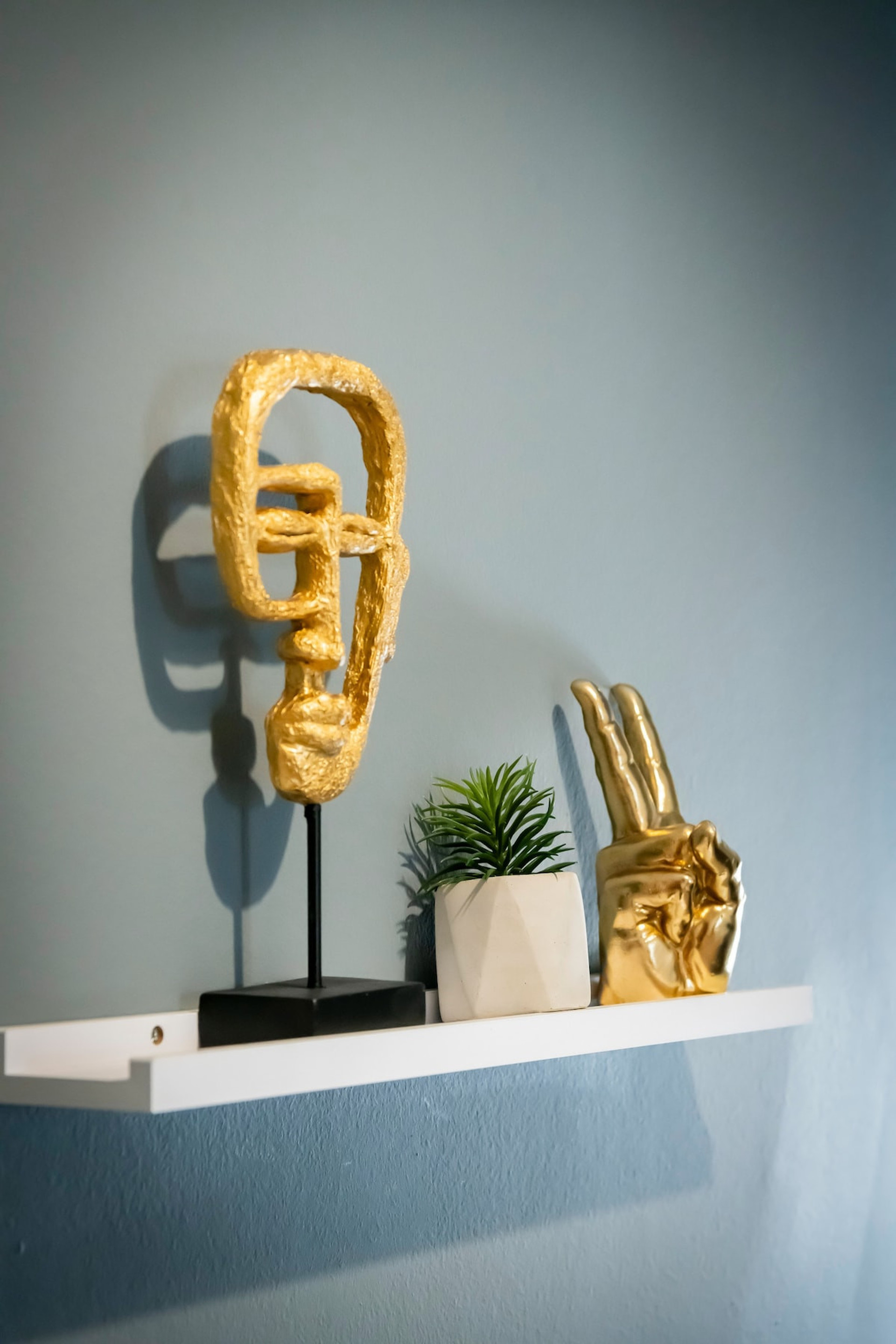 A stylish and modern detail: a sleek white shelf adorned with a golden abstract sculpture, a vibrant green plant in a chic pot, and a playful golden hand decor. Set against a cool blue wall, this adds charm and personality to the space.