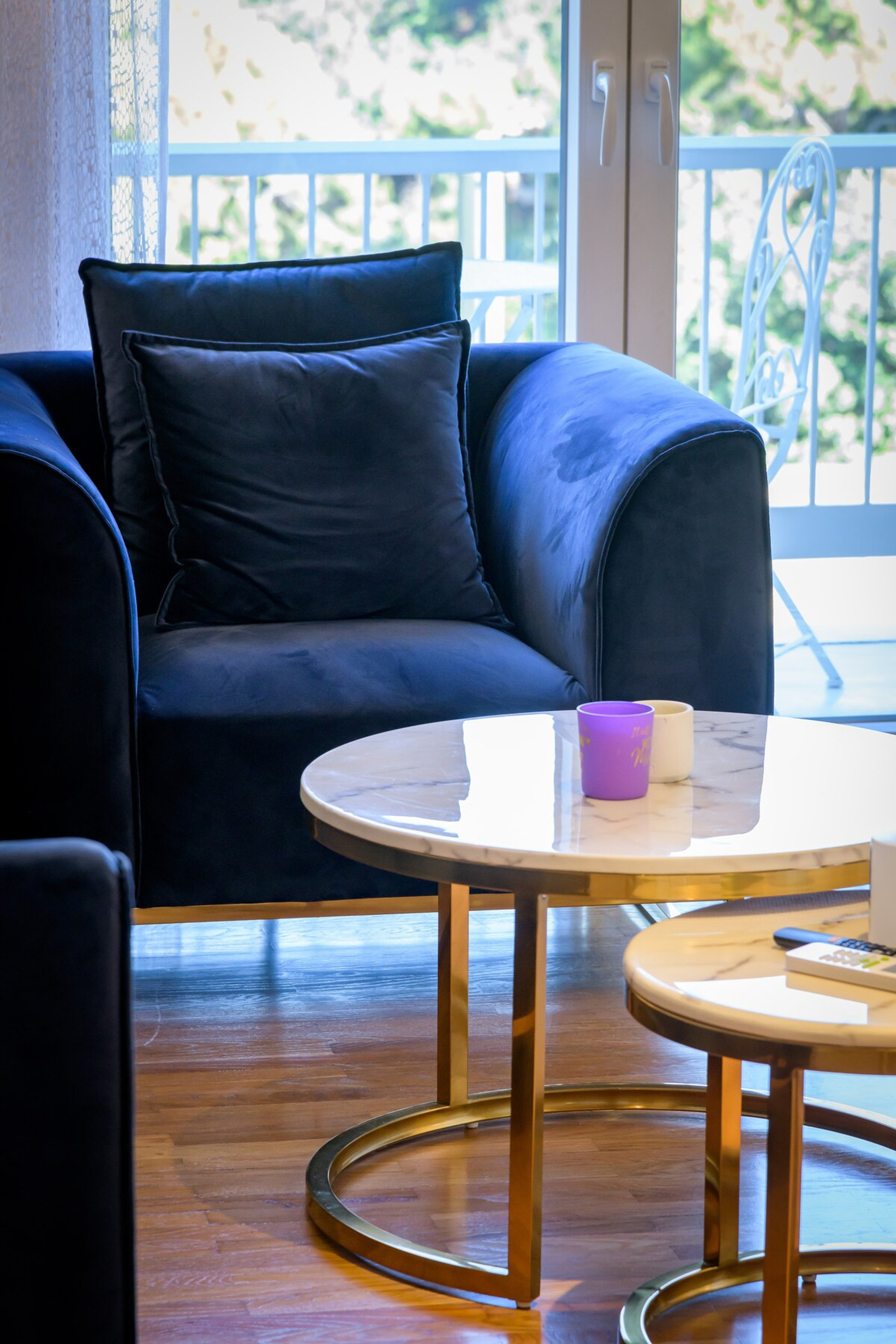 Cosy and stylish living space featuring a plush navy armchair, elegant gold-accented coffee tables, and a backdrop of natural light streaming through large windows with a view of greenery. Perfect for relaxing or enjoying a cup of coffee.