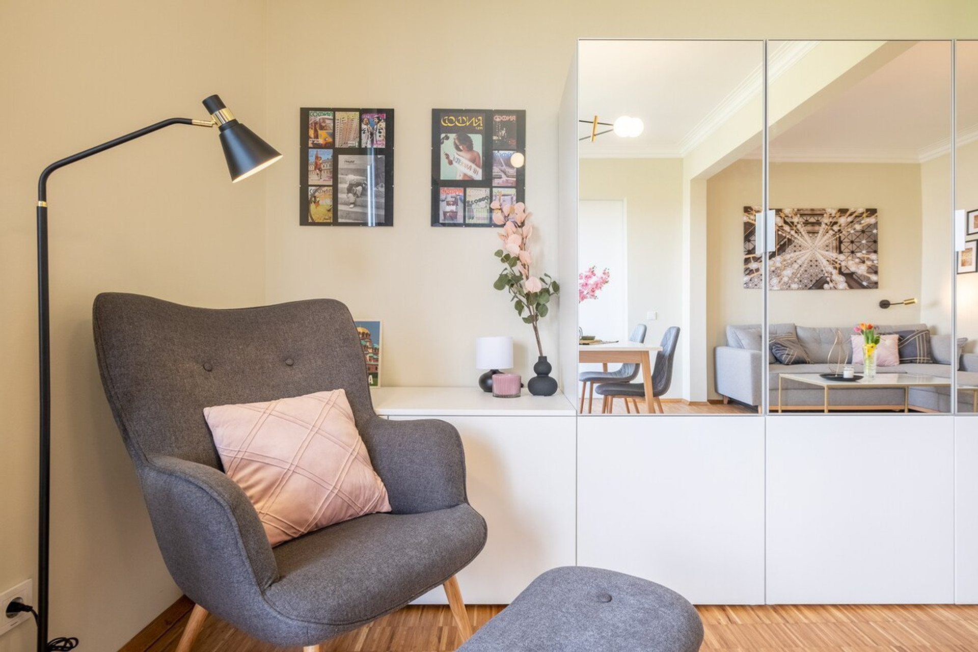 Cozy reading nook with a stylish armchair, blush pink pillow, and matching footrest. The corner is complemented by a sleek floor lamp and a white mirrored storage unit, reflecting the bright, open living space. A perfect spot to unwind with a book.