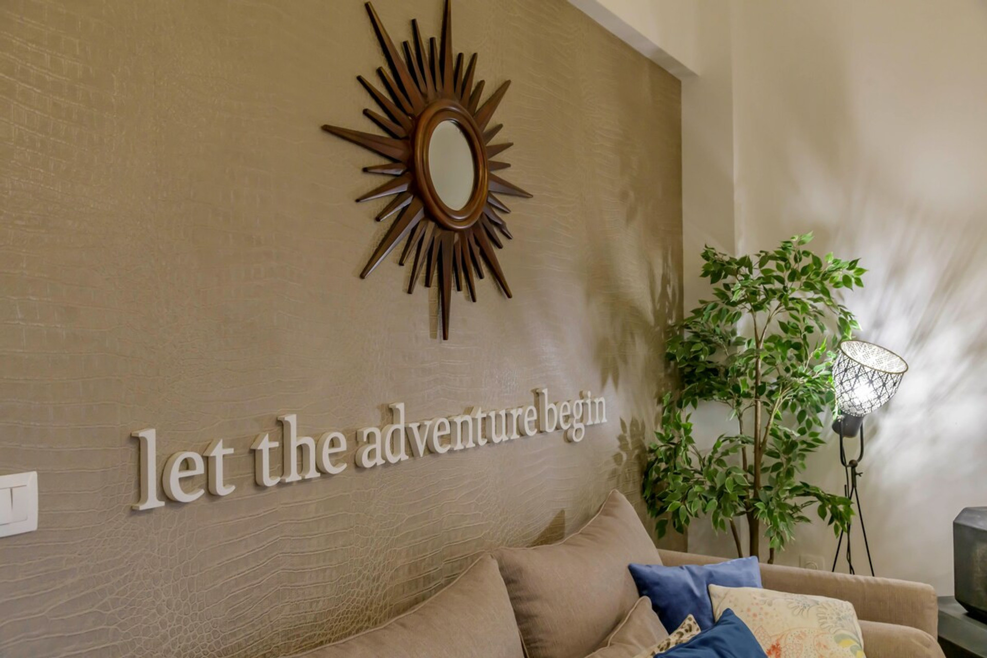 Cosy and inviting living space with a chic sunburst mirror, lush greenery, and a soft glow from elegant lighting – perfectly complemented by the inspiring "let the adventure begin" wall feature.