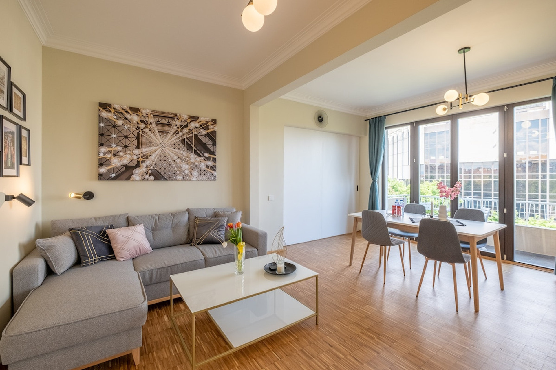 Super bright living room with stunning sunrise views overlooking NDK (National Palace of Culture). Features a cozy sectional sofa, warm wooden flooring, modern decor, and a dining table for four, perfect for meals/ work while enjoying natural light.
