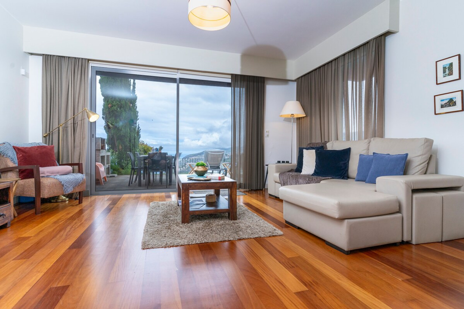 Modern living room with wooden floor, beige sofa with blue cushions, coffee table, armchair, and balcony overlooking the sea and mountains in the background.