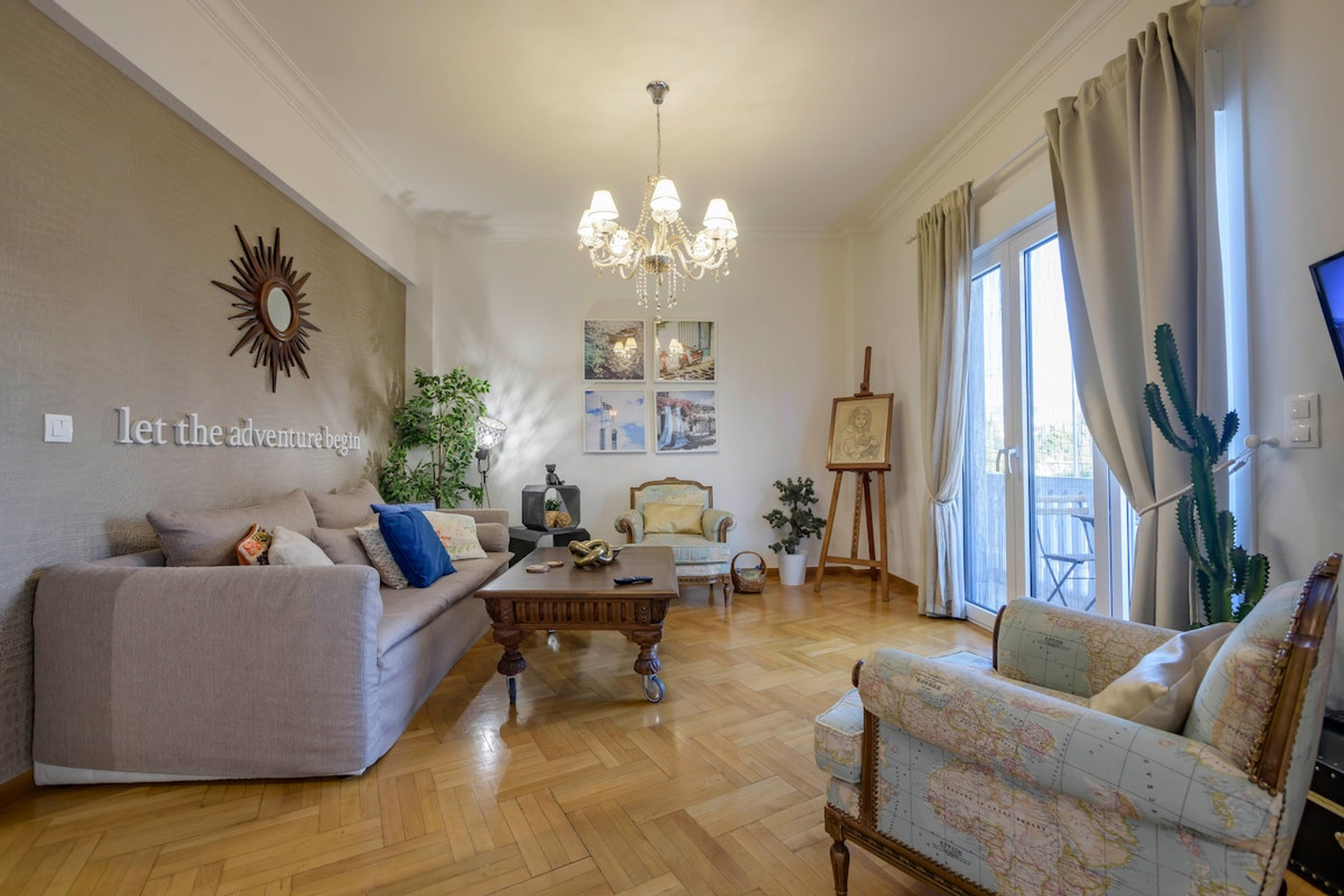 Bright and inviting living room featuring elegant decor, a cosy sofa, charming artwork, and a chandelier. Natural light pours in through large windows, offering a serene ambiance perfect for relaxation or unwinding with a book.