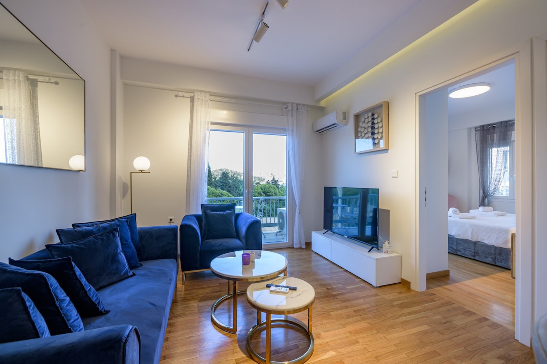 Bright and inviting living room featuring plush navy sofas, a smart TV, and chic gold-accented coffee tables. Natural light pours in through large windows, offering a serene view and seamless access to the cosy adjacent bedroom.