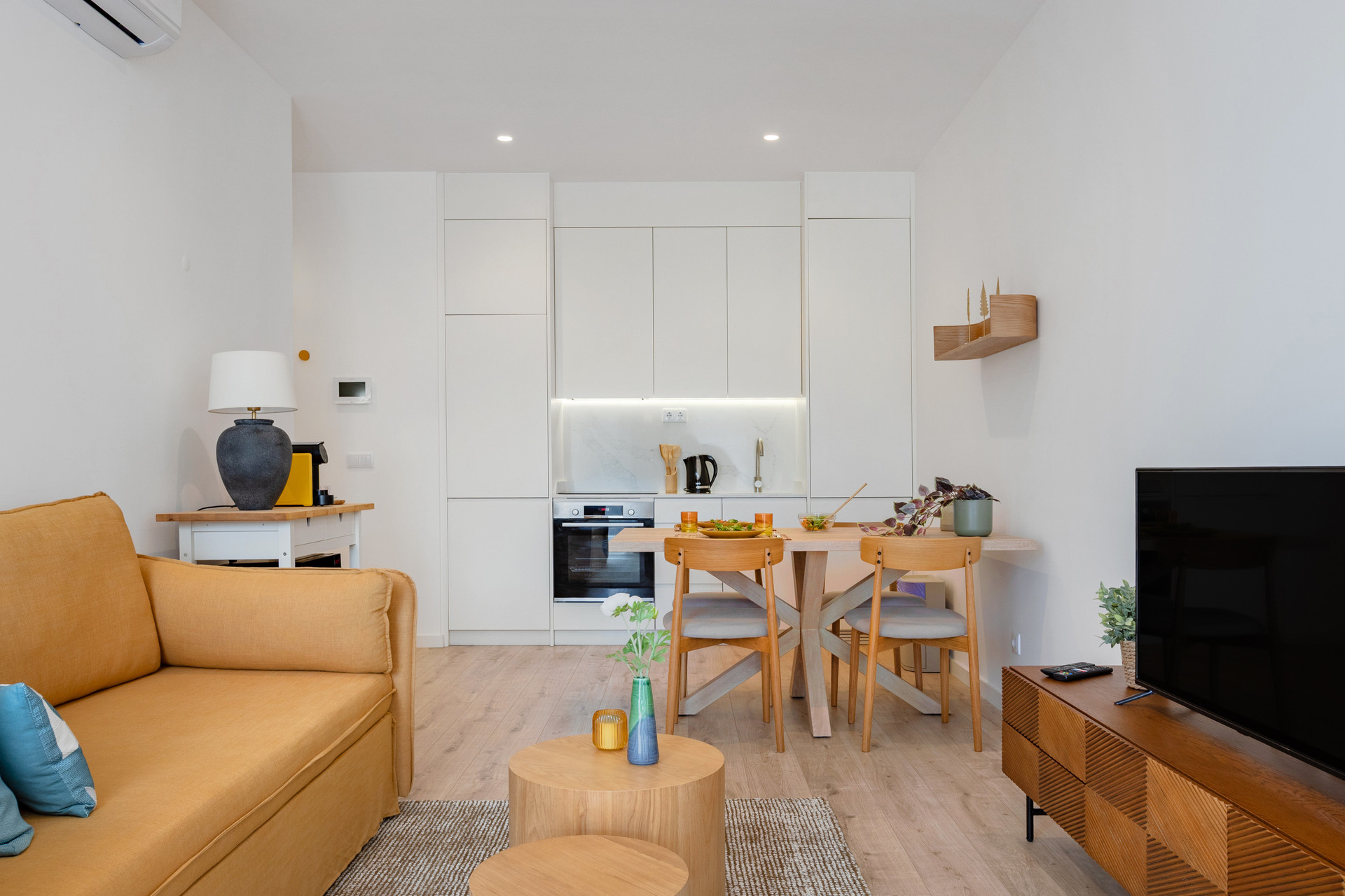 An open-plan layout filled with natural light and modern wood accents, featuring a cozy two-seater sofa, TV, and air conditioning.