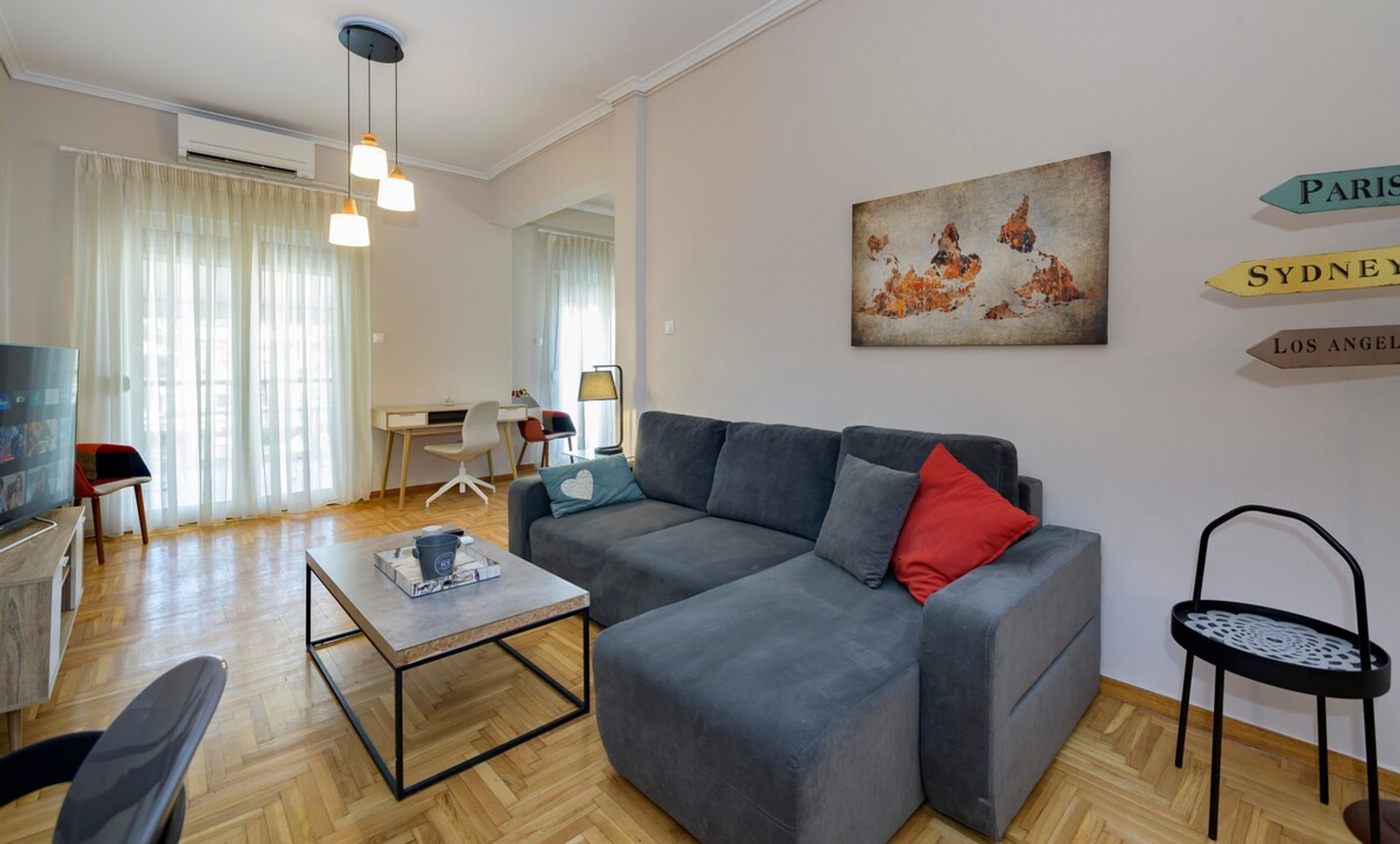 Bright and inviting living room featuring a cosy grey sofa adorned with vibrant cushions, a stylish coffee table, and a smart TV. Natural light pours in through large windows, complemented by tasteful decor and a charming workspace for added comfort.