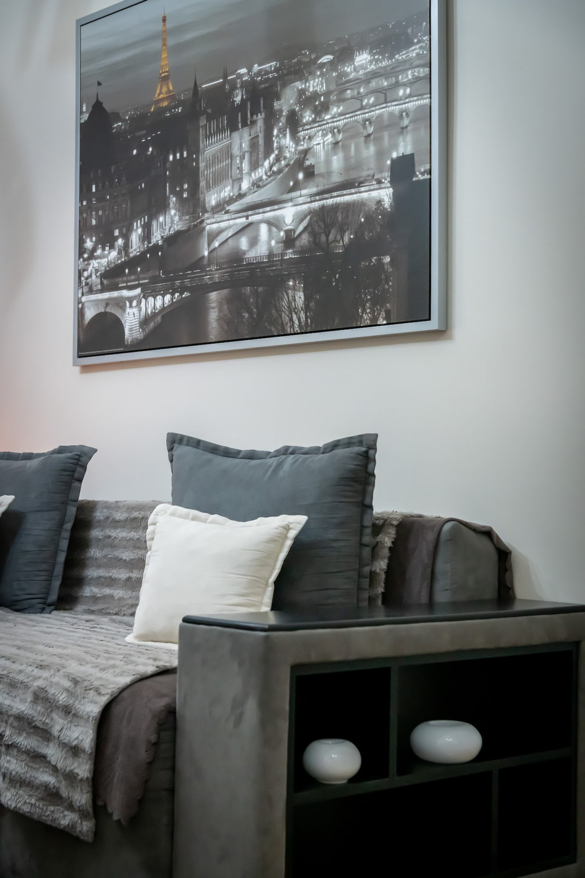 Cosy and stylish living space with a plush sofa adorned with soft cushions and a throw, complemented by a chic side table. A stunning Parisian cityscape artwork adds an elegant touch, creating a warm and inviting atmosphere. Perfect for relaxation.