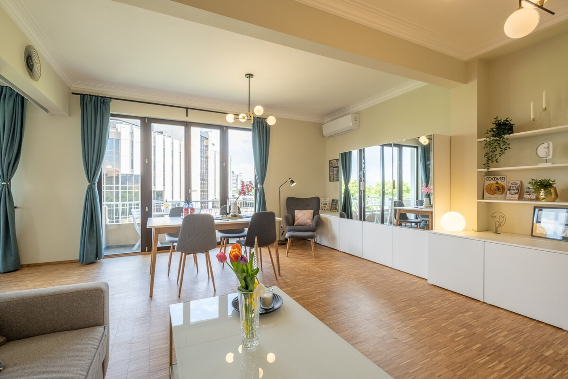 Modern open-plan living area with a cozy sofa, a dining table for four, and large windows for natural light. The mirrored storage unit adds to the spaciousness, while the balcony offers outdoor relaxation and views of NDK.