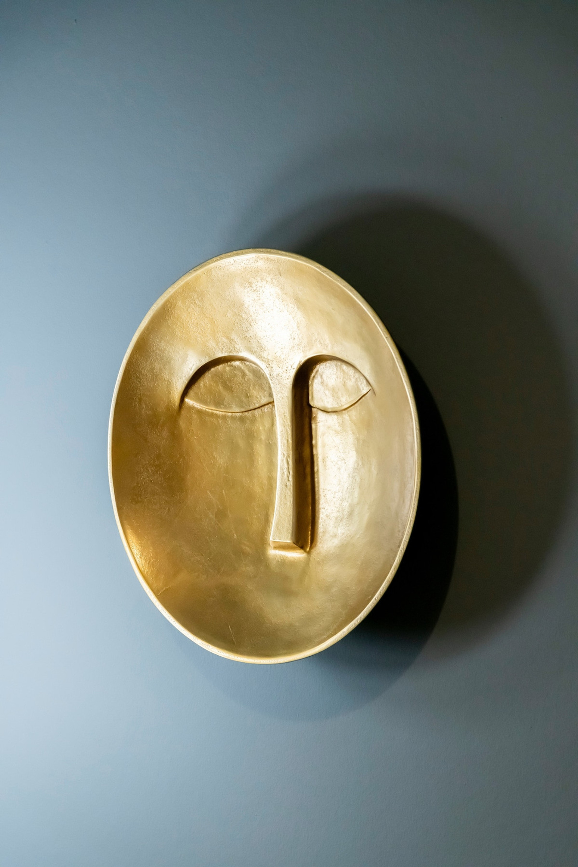 A striking golden wall sculpture with a minimalist, abstract face design, set against a sophisticated grey backdrop. Perfect for adding an artistic touch and a sense of refined elegance to the living space.