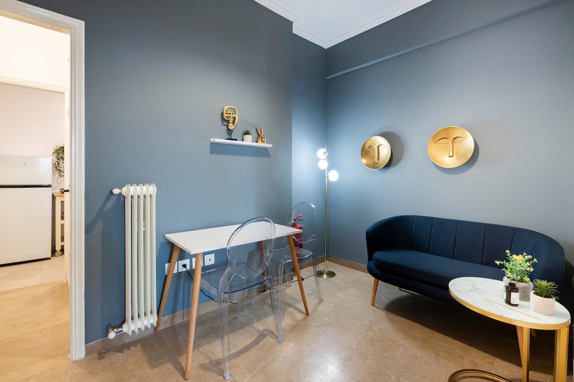 Cosy and stylish living room featuring a modern navy sofa, a chic glass desk, and warm lighting accents. The elegant decor, including gold wall art and vibrant greenery, creates a welcoming atmosphere perfect for relaxation or productivity.